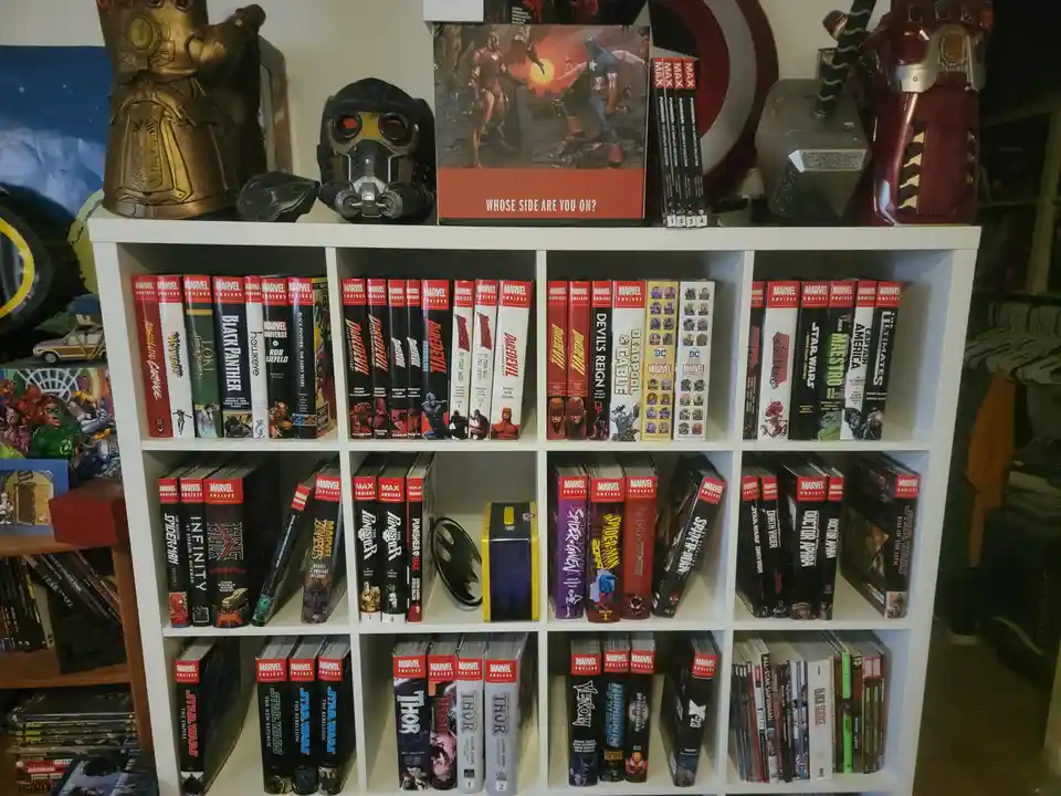 A white bookshelf filled with Marvel and Star Wars comic books, topped with superhero memorabilia including a Thanos…