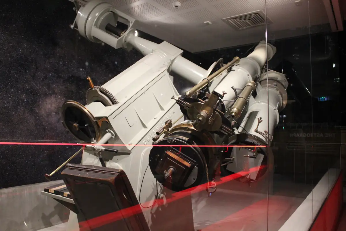 A large, white, vintage astronomical telescope is displayed behind glass in a museum, with a red laser line cutting…