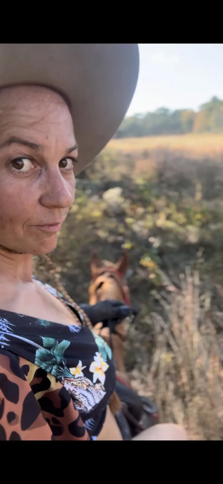 A woman wearing a cowboy hat and a colorful patterned top looks directly at the camera with a slightly skeptical or…
