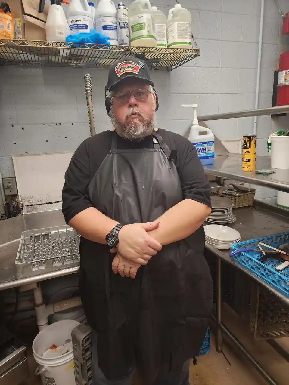 A man with a beard and glasses, wearing a black apron and headphones, stands in a commercial kitchen with arms crossed…