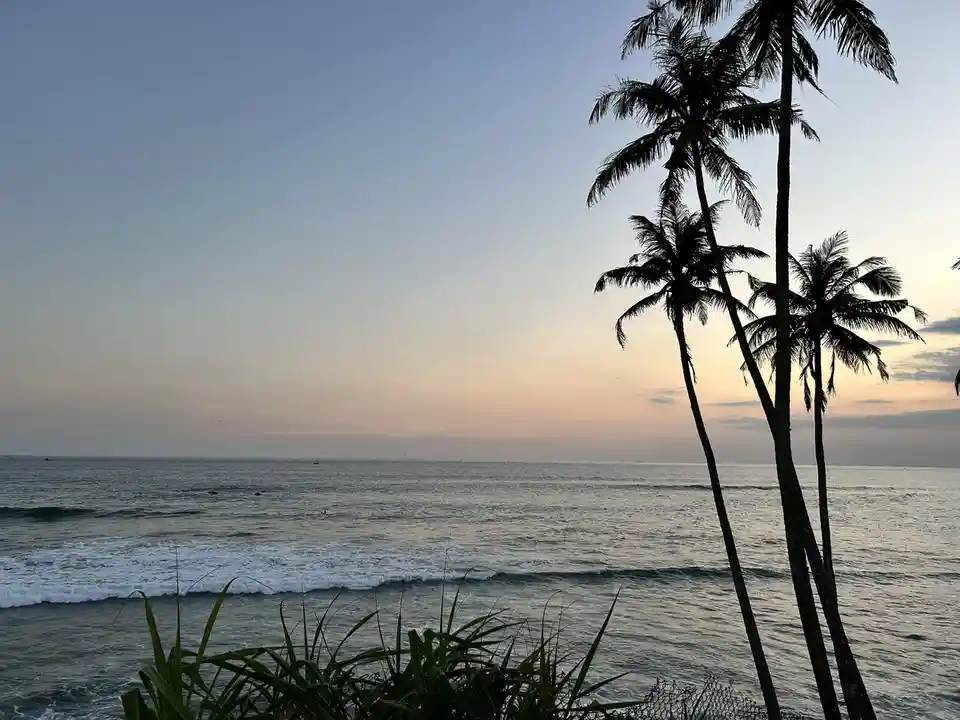A serene tropical beach scene at sunset, featuring silhouetted palm trees against a gradient sky transitioning from blue to…
