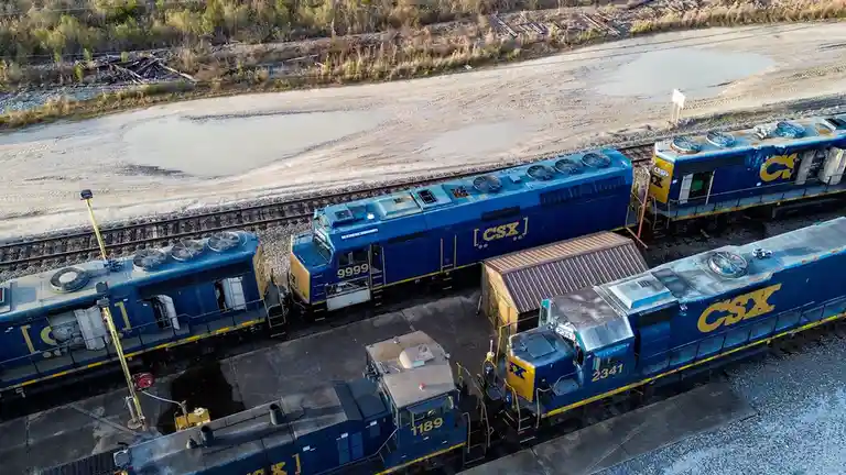An aerial view shows four blue CSX locomotives parked on adjacent tracks in a rail yard, with one engine prominently…