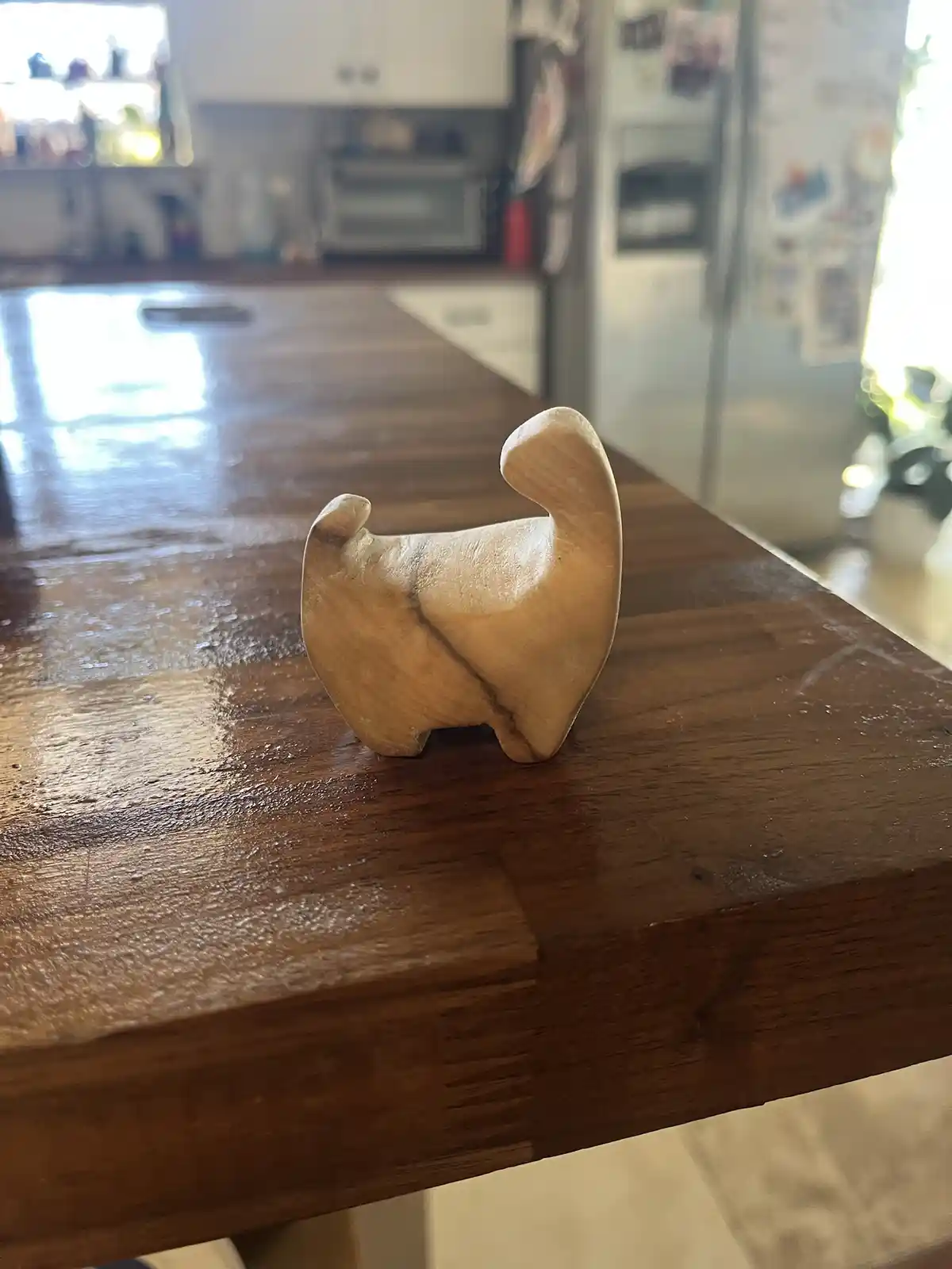 A simple wooden carving of a dog, viewed from behind, stands on a polished wooden kitchen counter. The carving has a…