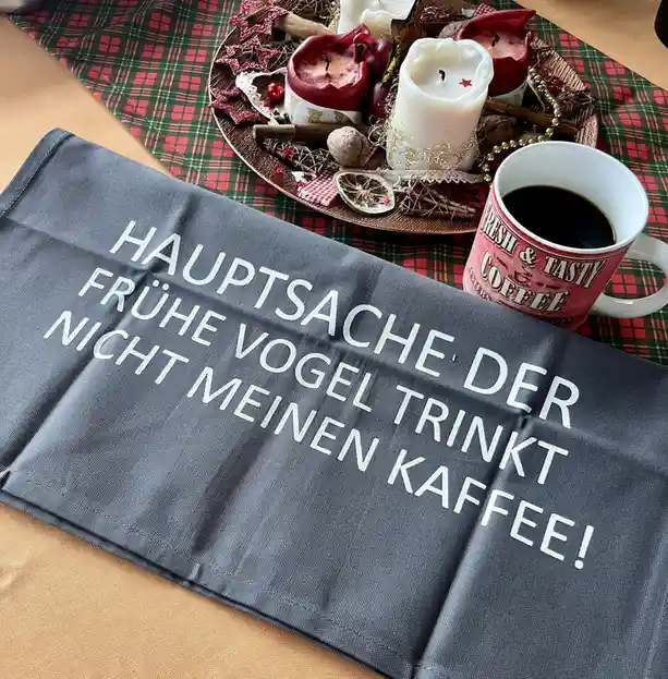 A grey cloth with white text reading 'HAUPTSACHE DER FRÜHE VOGEL TRINKT NICHT MEINEN KAFFEE!' is placed on a wooden table…