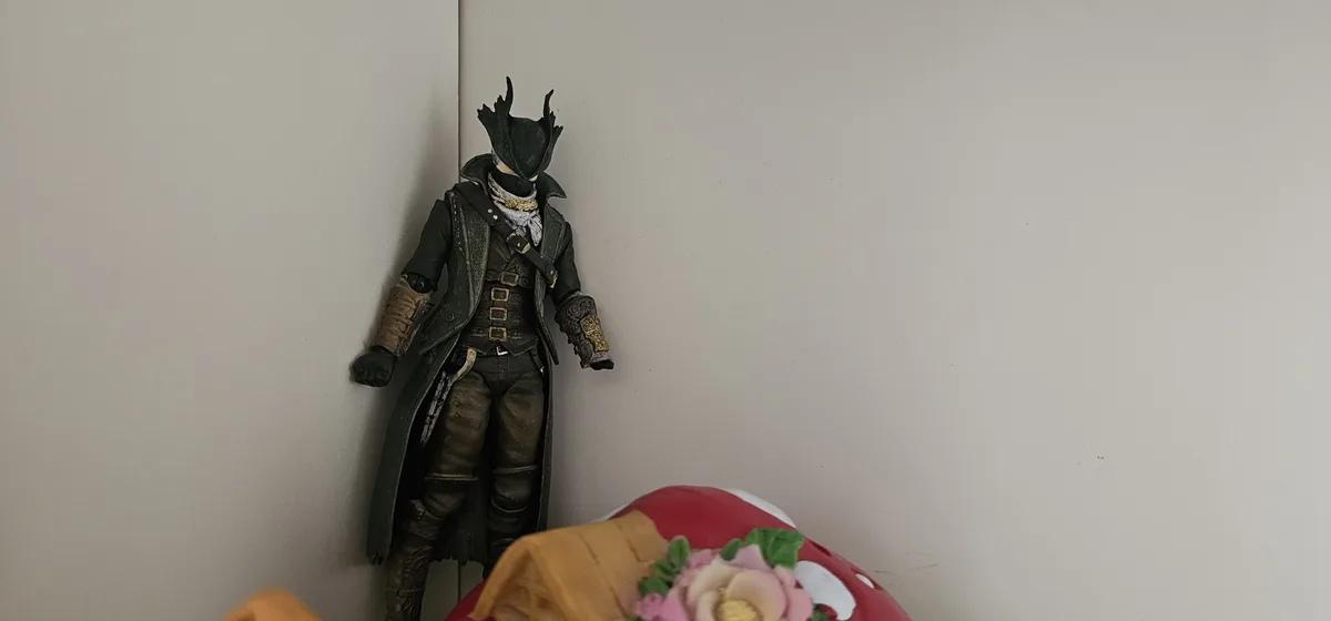 A darkly dressed action figure with a horned mask and long coat leans against a plain wall, positioned above a partially…