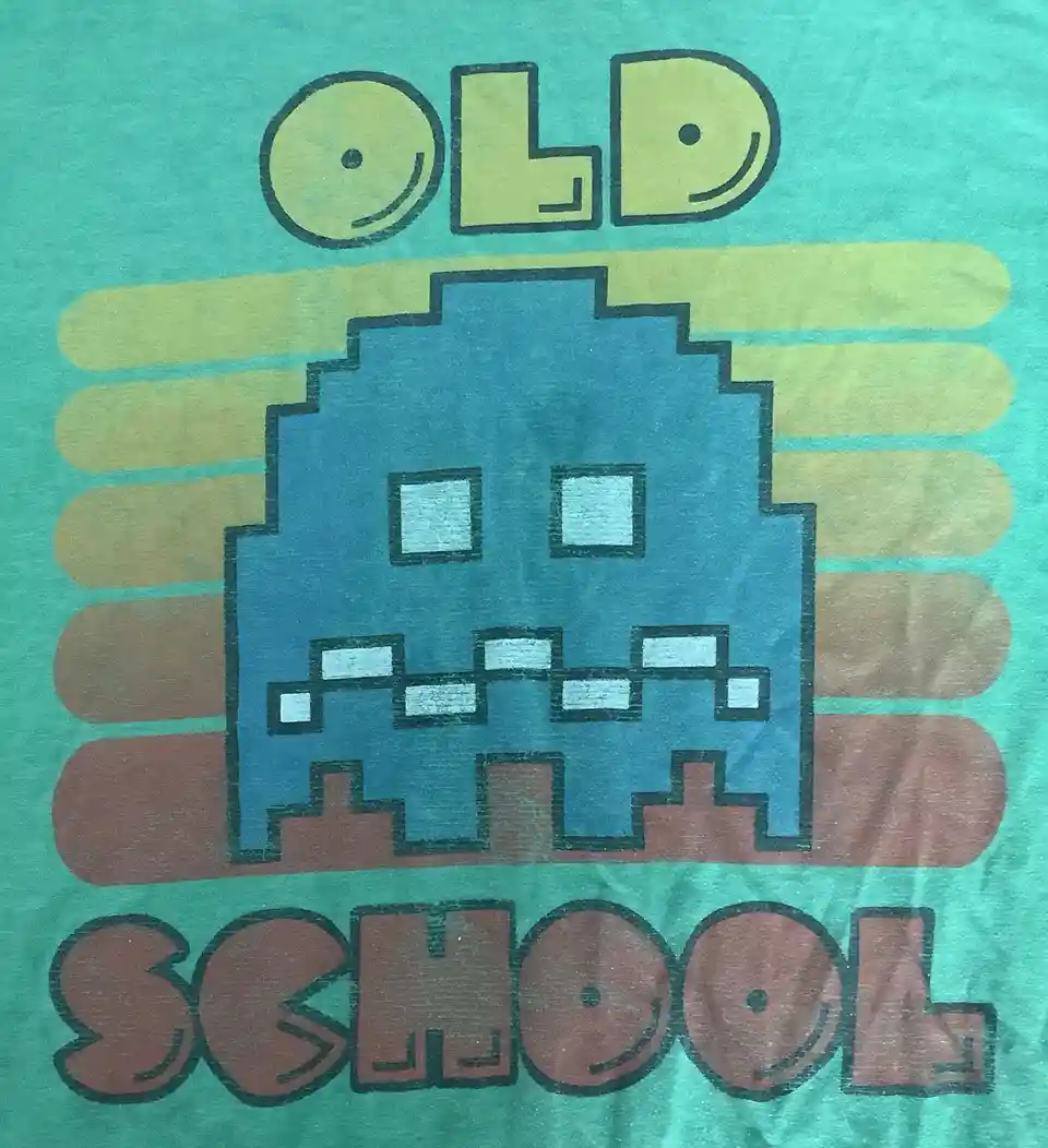 A close-up of a teal t-shirt featuring a pixelated blue alien character with square eyes and teeth, set against a background…