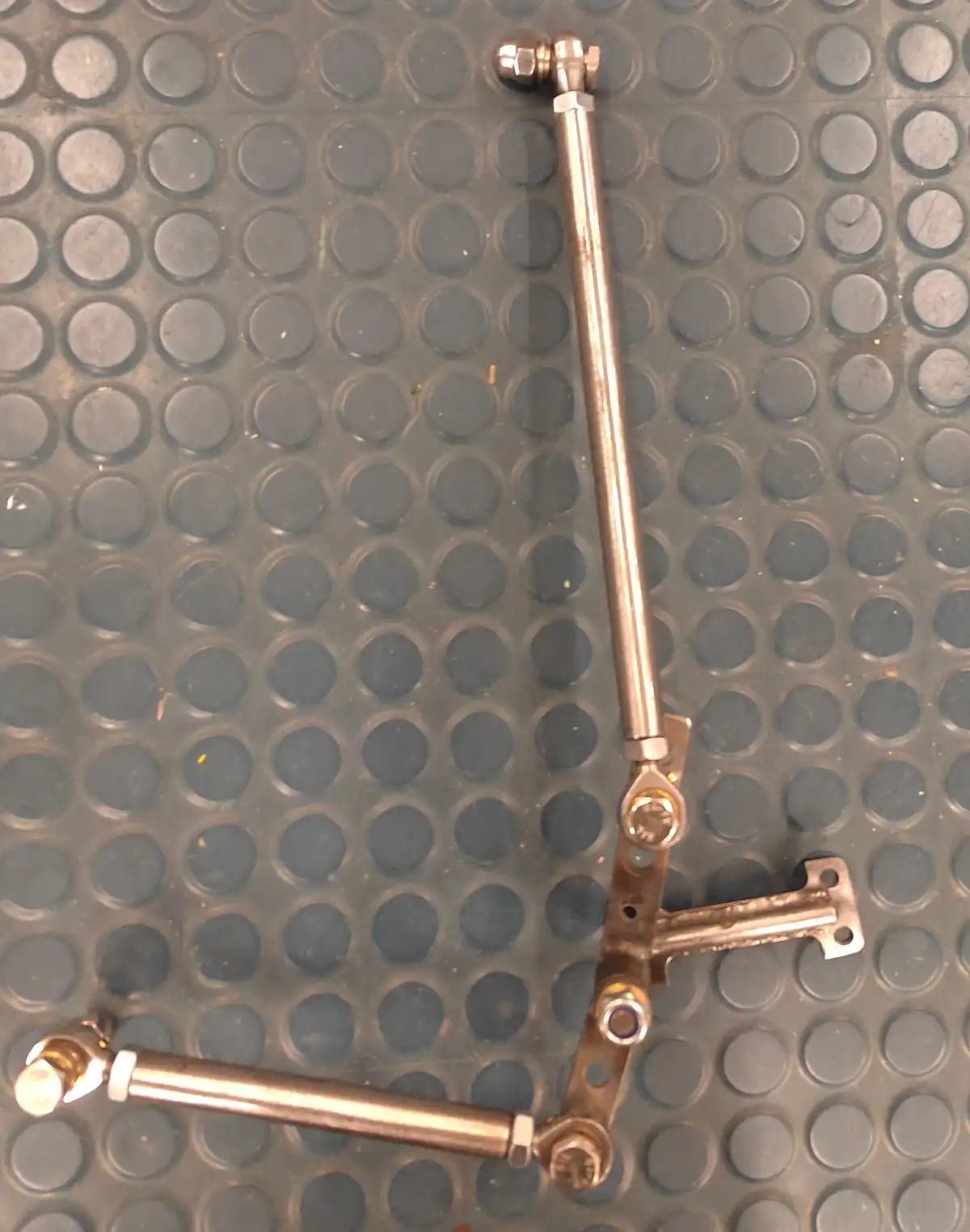 A metal mechanical linkage with two rods and a bracket lies on a gray, textured, circular-patterned floor. The overlaid text…