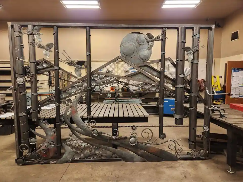 A large, ornate metal gate made of welded steel pipes, decorative swirls, and spherical elements sits in a workshop. The…