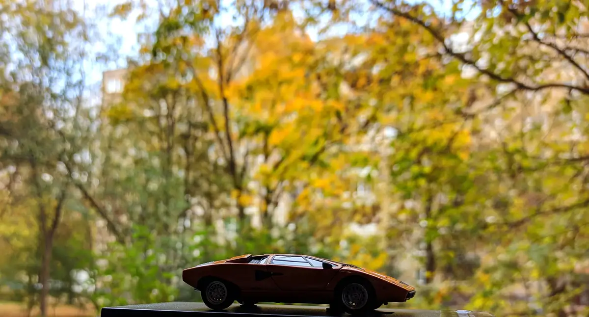 A small, brown die-cast model of a Lamborghini Countach is shown in sharp focus on a black surface, with a blurred…