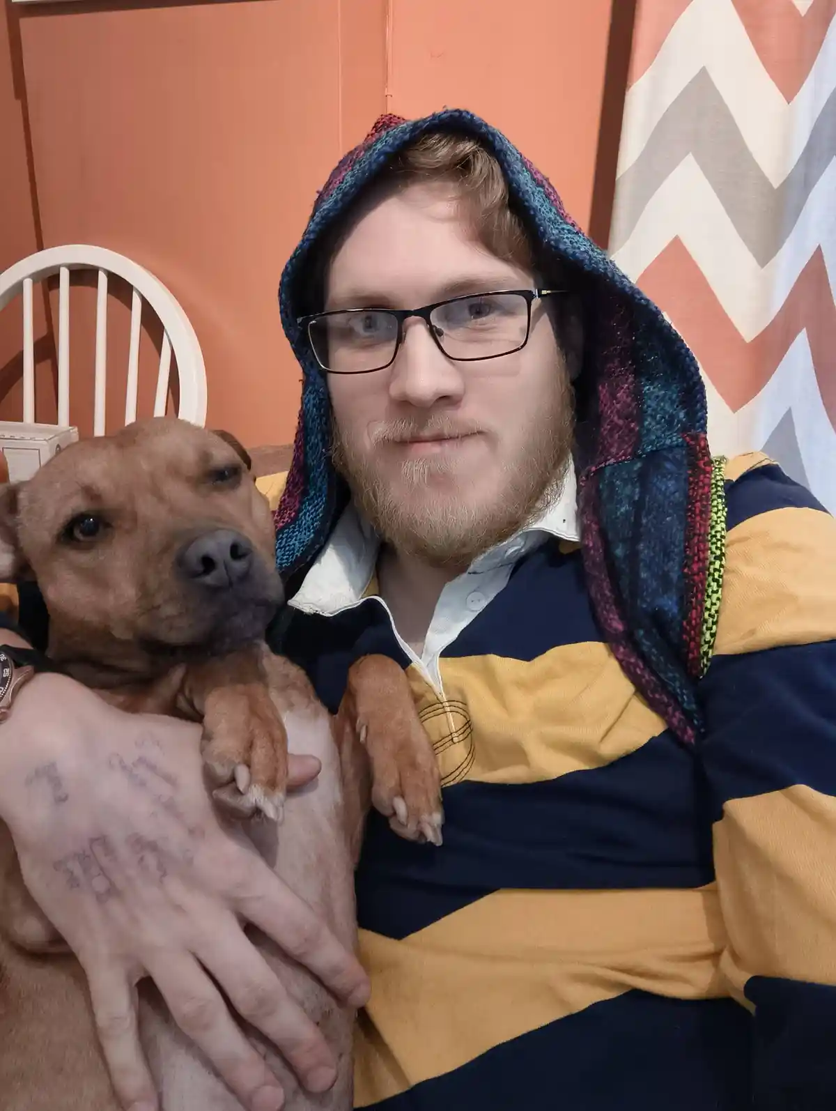 A man with glasses and a colorful hooded blanket over his head is holding a brown dog. The dog has its paws resting on the…