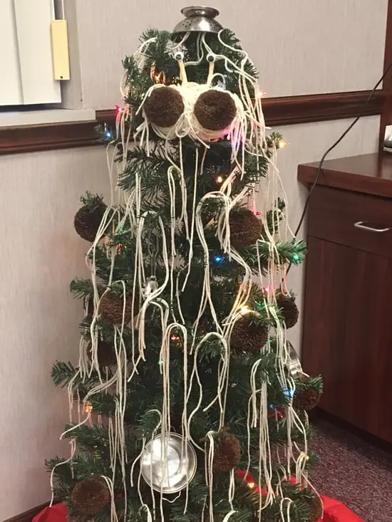 A decorated christmas tree is covered in white yarn, brown pom-poms, and lights, with a metal pot on top and a yarn 'face'…