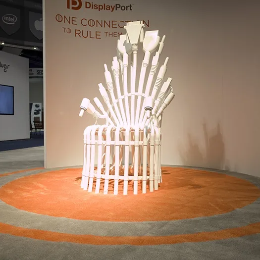 A white throne made of DisplayPort cables sits on an orange circular rug at a tech expo. Behind it, a wall displays the…