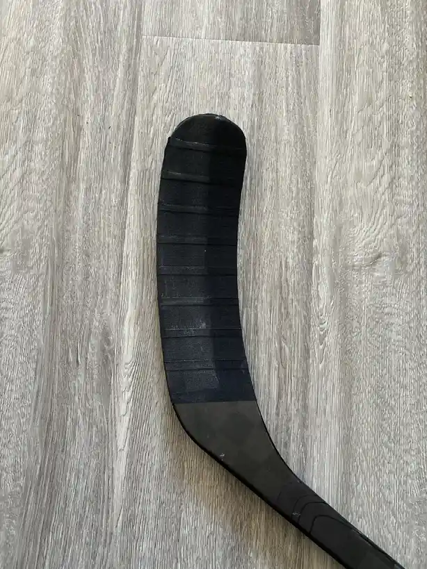 A close-up photo shows the blade of a black hockey stick with textured grip tape lying on a light gray wood-grain floor. The…