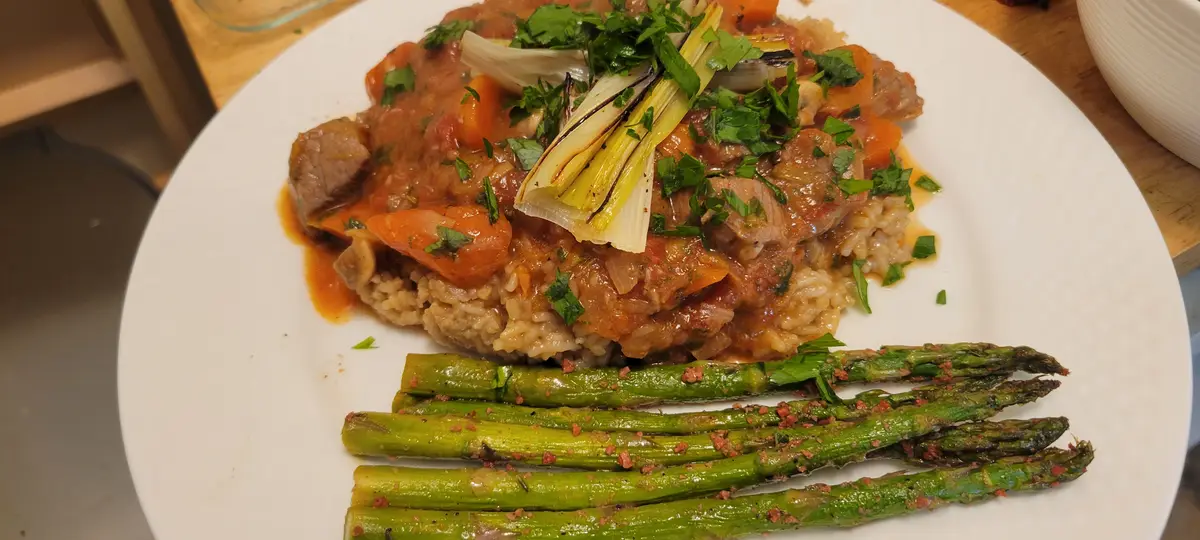 A close-up photo of a plated meal featuring a hearty stew with chunks of meat, carrots, and leeks over brown rice, garnished…