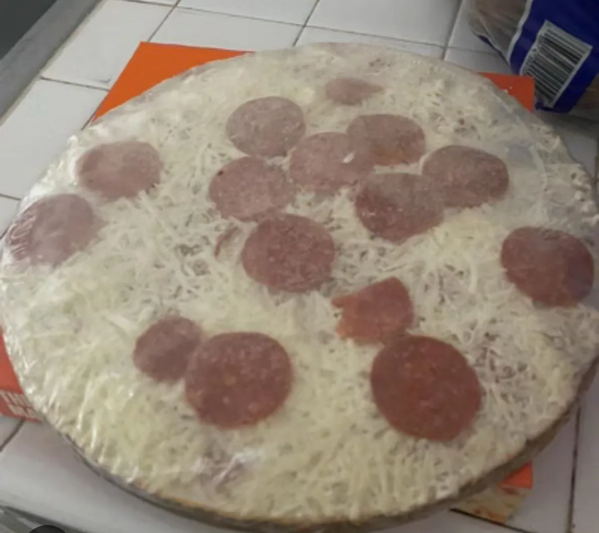 A round pizza with shredded cheese and pepperoni slices is completely covered in plastic wrap, sitting on an orange…