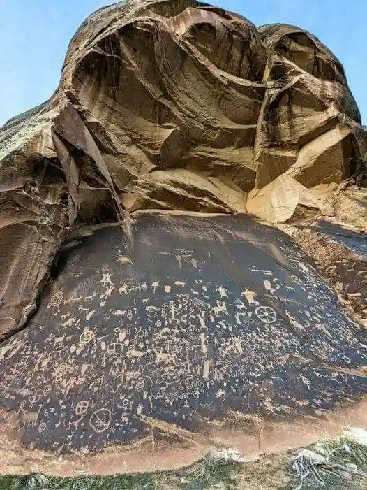 A large, weathered rock face covered in numerous ancient petroglyphs, including human figures, animals, and geometric shapes…