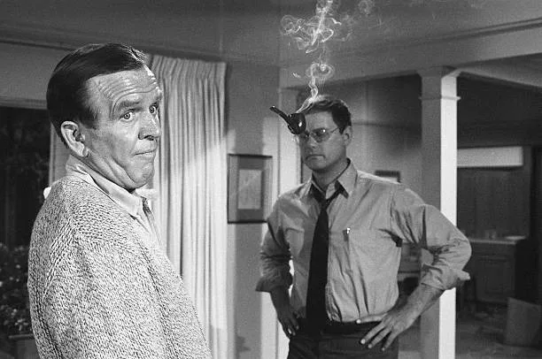 A black-and-white still from a 1960s TV show shows two men in a room. The man on the left, in a cardigan, looks surprised…