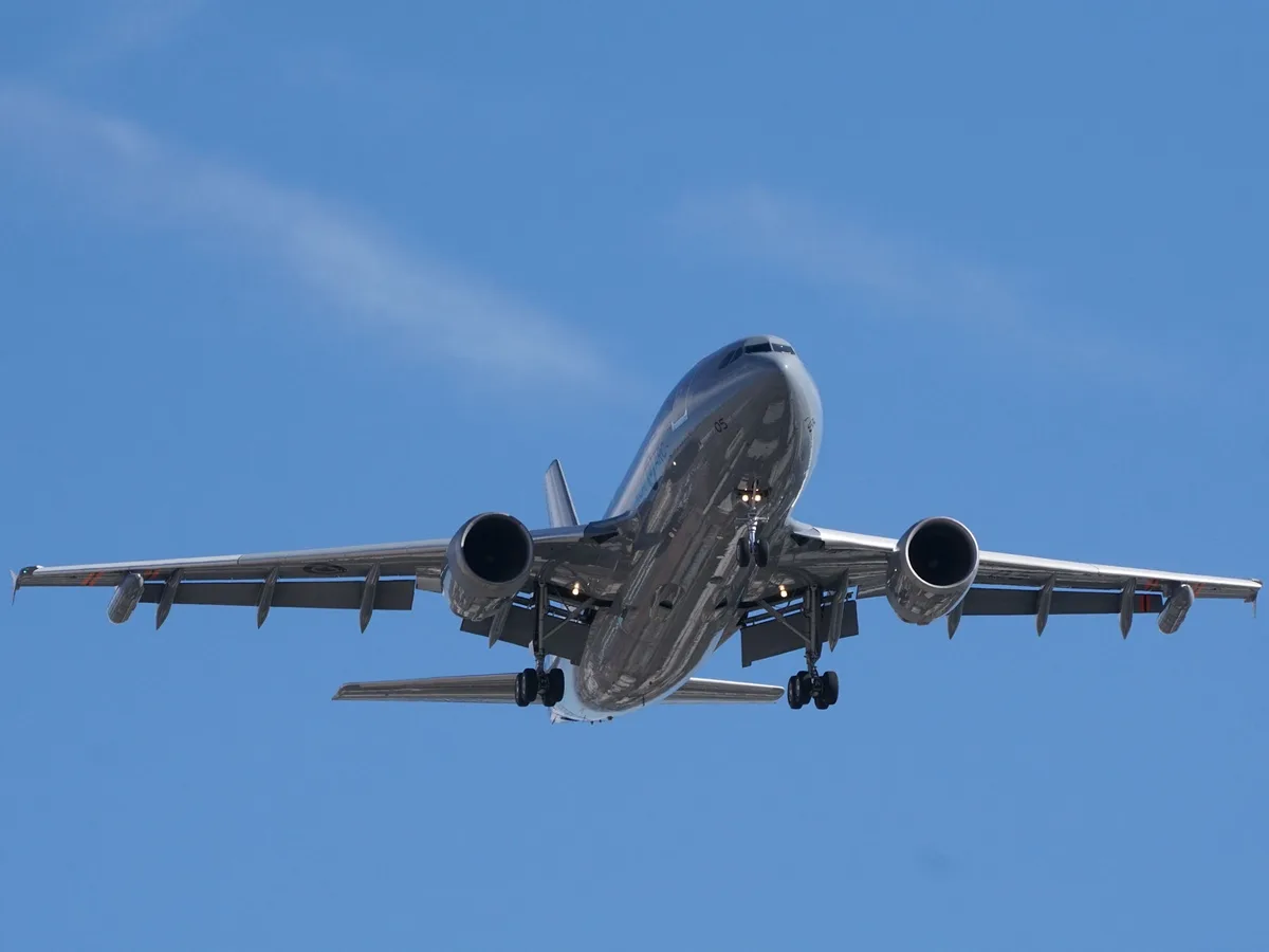 A large commercial airplane is captured from below, flying against a clear blue sky with its landing gear down. The…