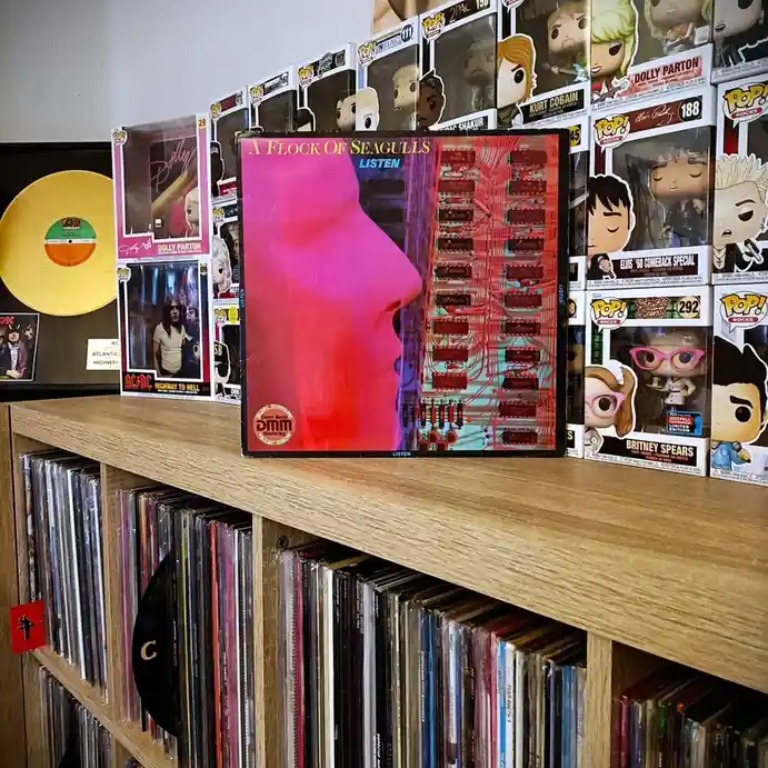 A vinyl record album cover for 'A Flock of Seagulls - Listen' sits prominently on a wooden shelf, surrounded by Funko Pop!…