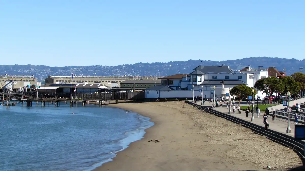 A wide shot of a sunny beachfront with calm water, a wooden pier, and buildings along the shore, with people walking on a…
