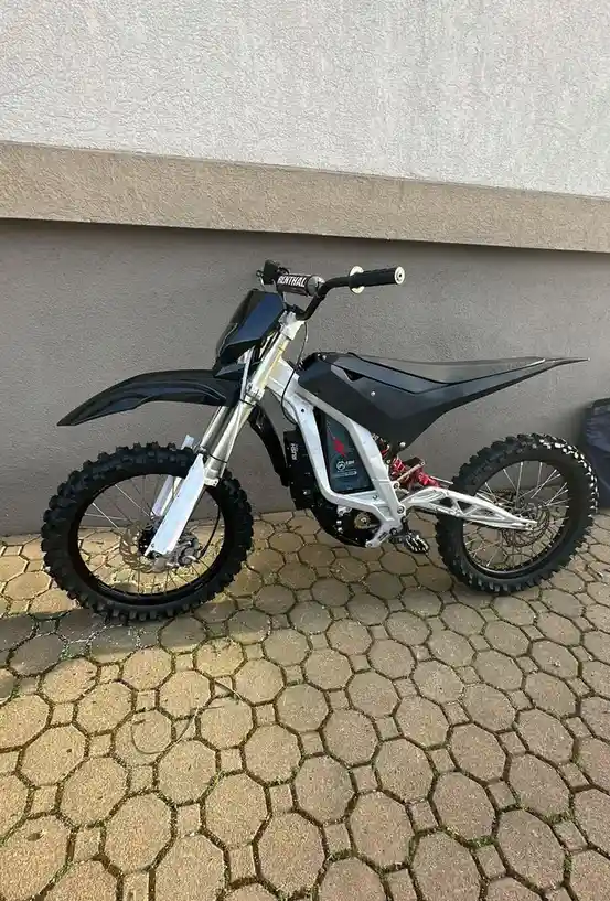 A silver and black electric dirt bike with knobby tires and red suspension is parked on a hexagonal stone patio against a…