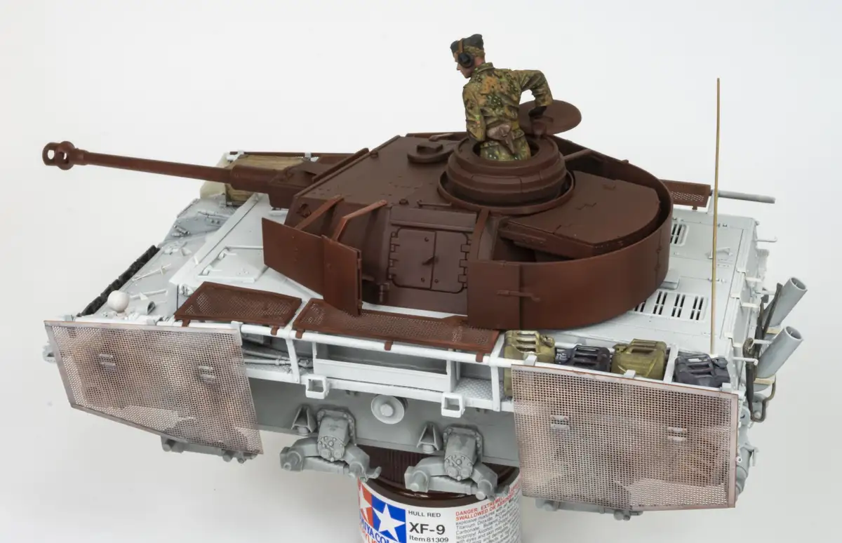 A detailed scale model of a tank with a miniature soldier figure in a camouflage uniform standing on its turret, looking…