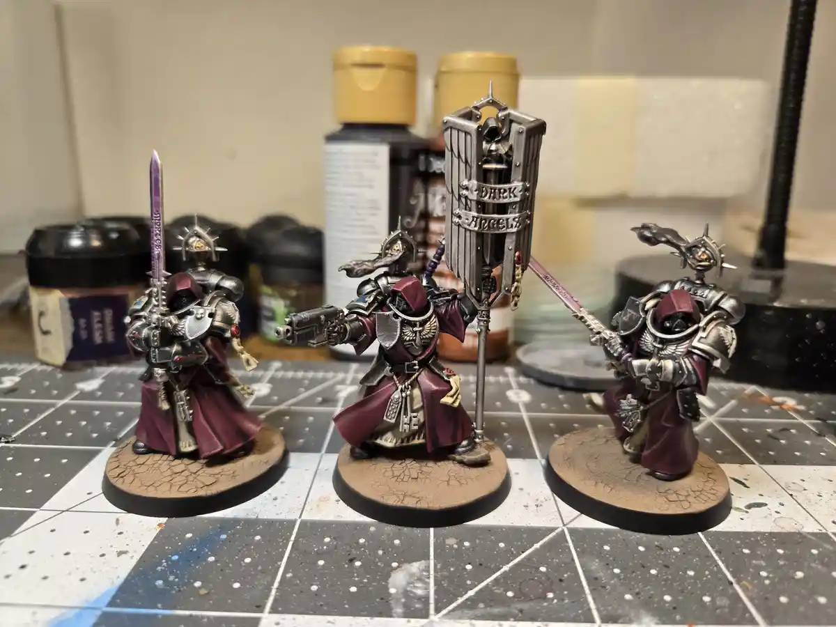 Three painted miniature Dark Angels Space Marines stand on circular bases on a gridded painting mat, surrounded by paint…