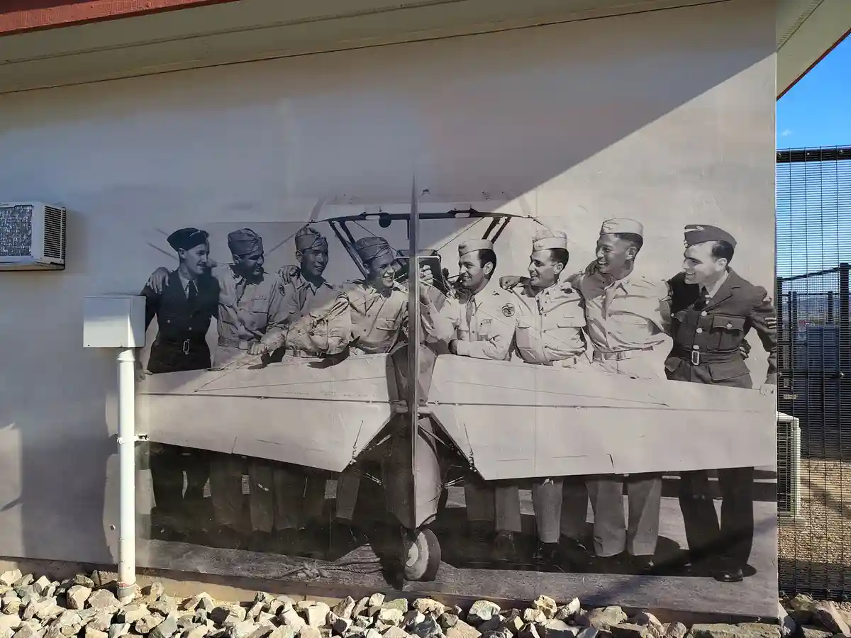 A black-and-white mural on a building wall depicts a group of smiling soldiers posing around a vintage biplane. The image is…