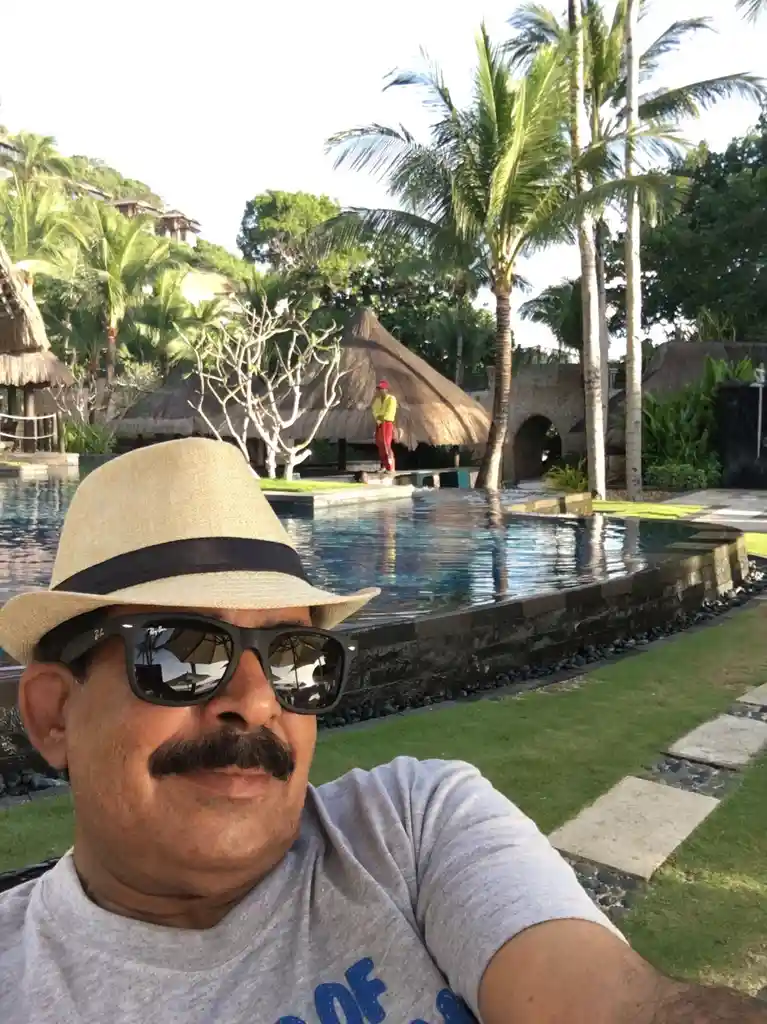 A man with a mustache, wearing a straw hat and sunglasses, takes a selfie in front of a tropical resort pool. In the…