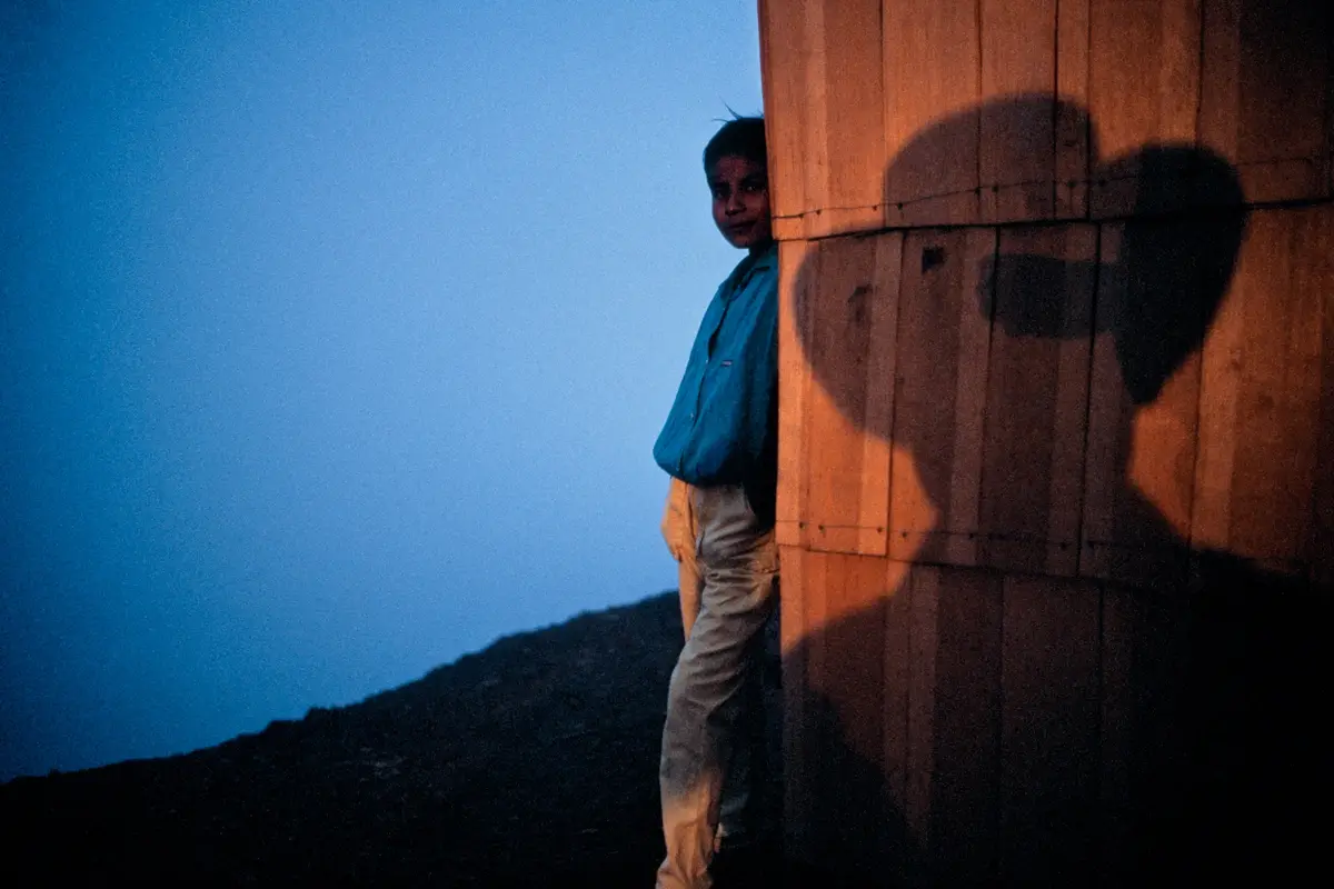 A young boy in a blue shirt and beige pants peeks from behind a wooden wall at dusk, his shadow cast on the wall. The shadow…