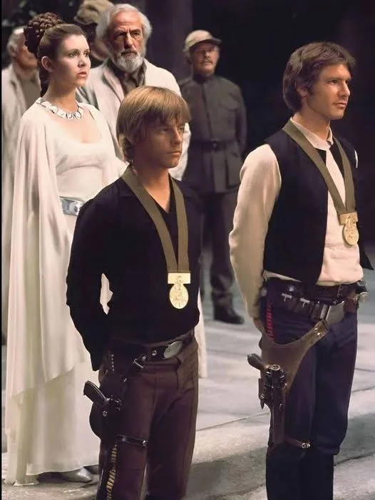 A still from Star Wars: Episode IV showing Luke Skywalker and Han Solo standing side-by-side, both wearing medals around…