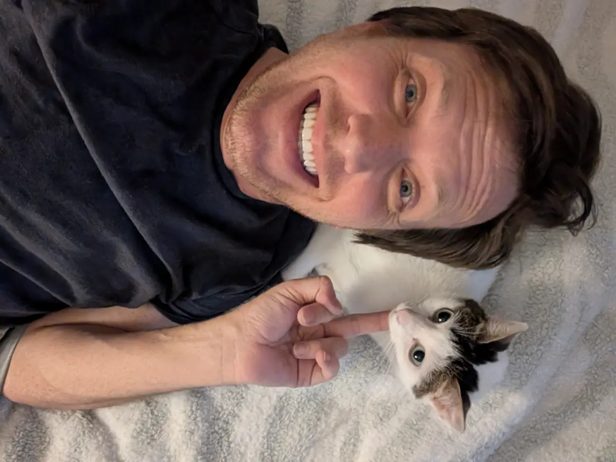 A man with brown hair and a black t-shirt lies on a grey blanket, grinning widely with his teeth showing while pointing his…