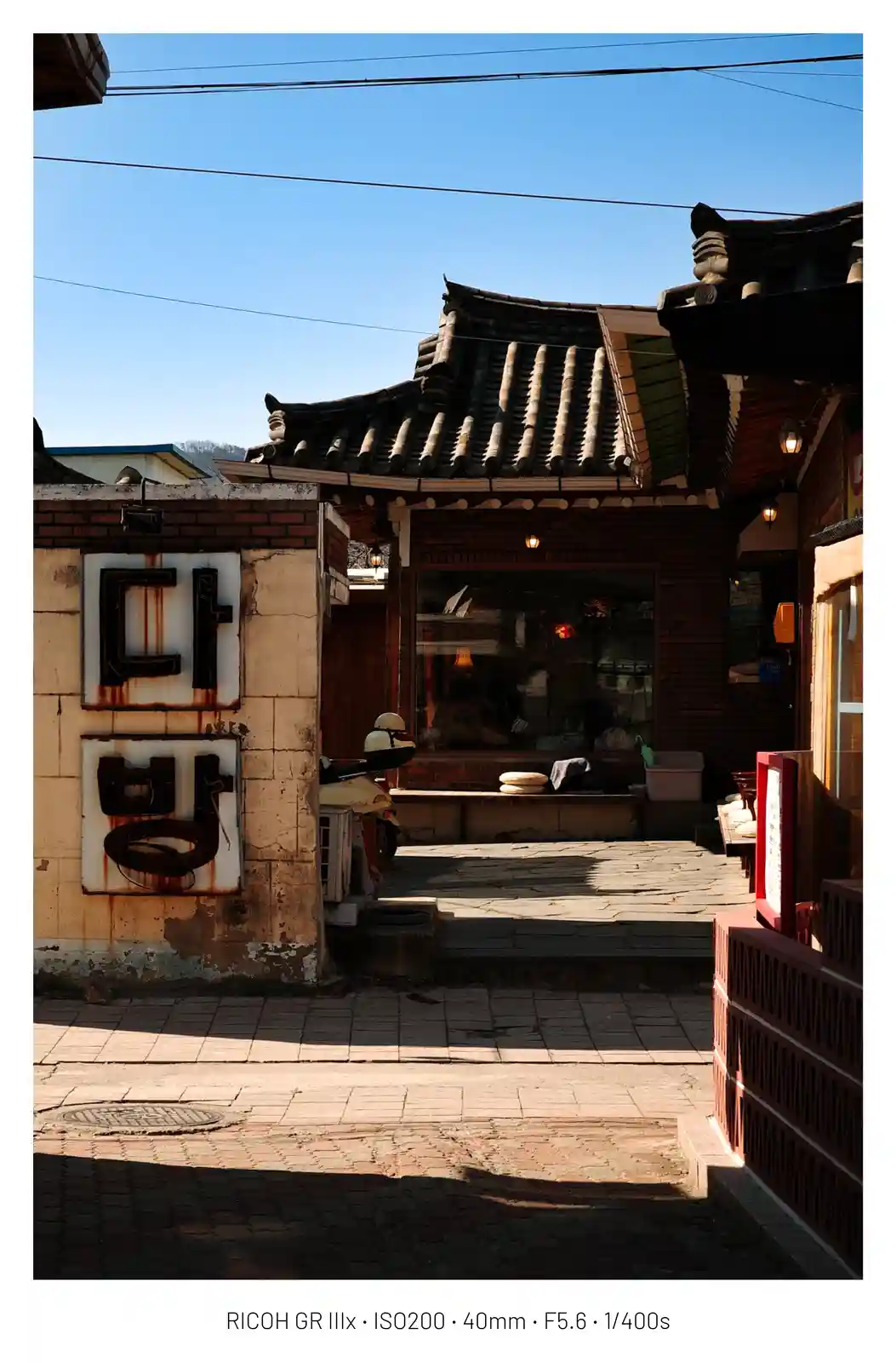 A photograph of a narrow alleyway leading to a traditional Korean building with a tiled roof, featuring a rusty metal sign…