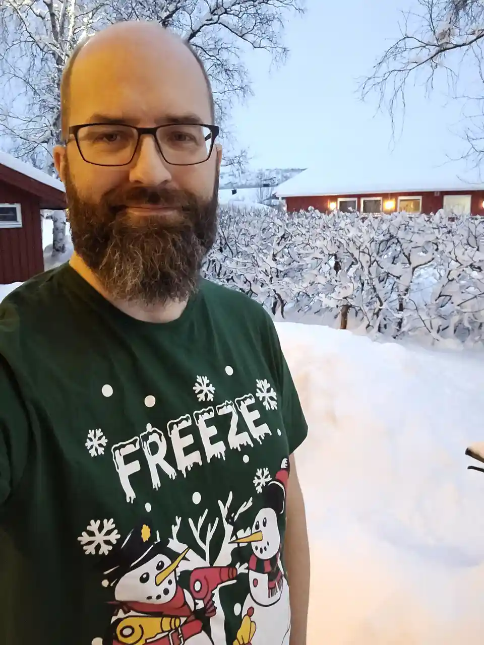 A man with glasses and a beard stands in a snowy outdoor setting, wearing a green t-shirt with snowmen and the word 'FREEZE'…