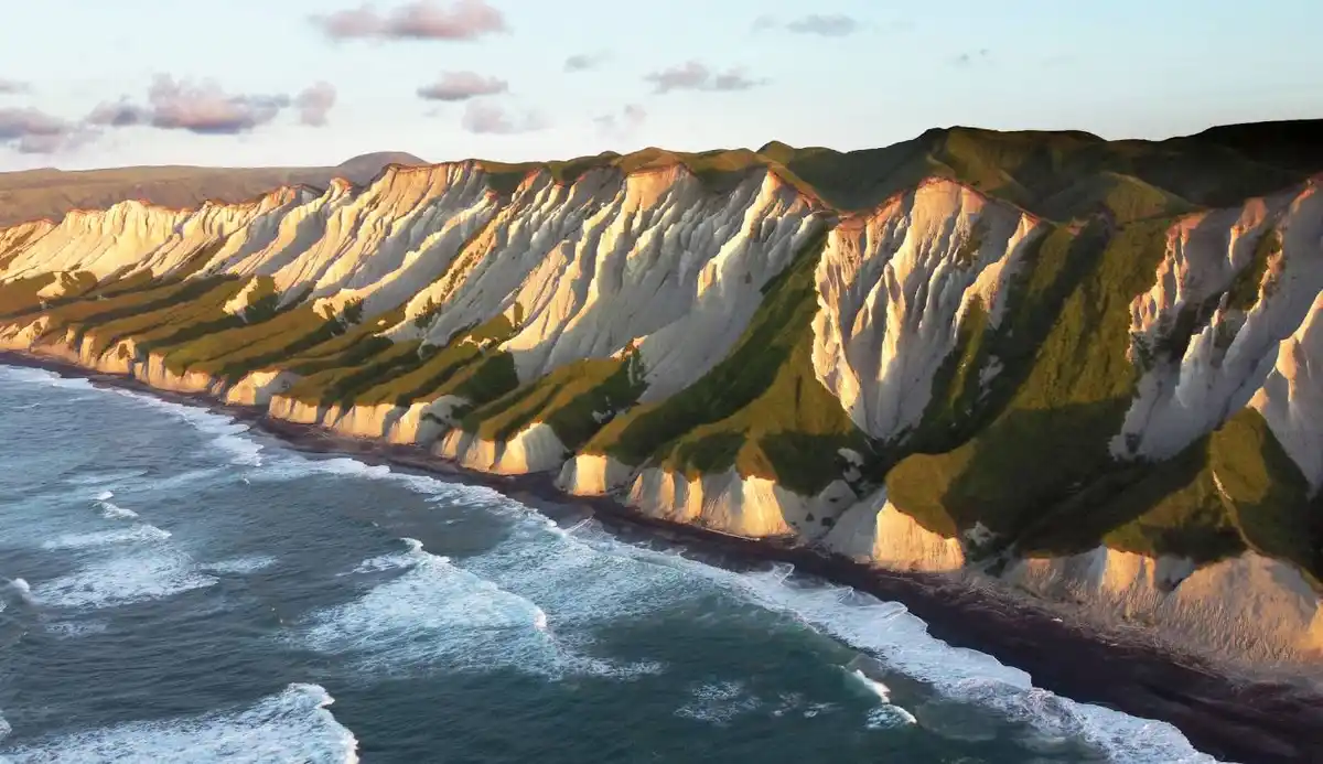Aerial view of dramatic white cliffs with green vegetation meeting a dark beach and ocean waves under a soft sky. Overlaid…