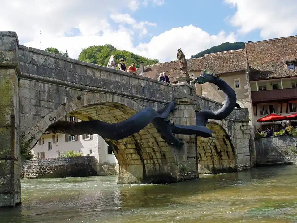 A large black dragon sculpture is positioned under a stone bridge spanning a river, with people standing on the bridge's…