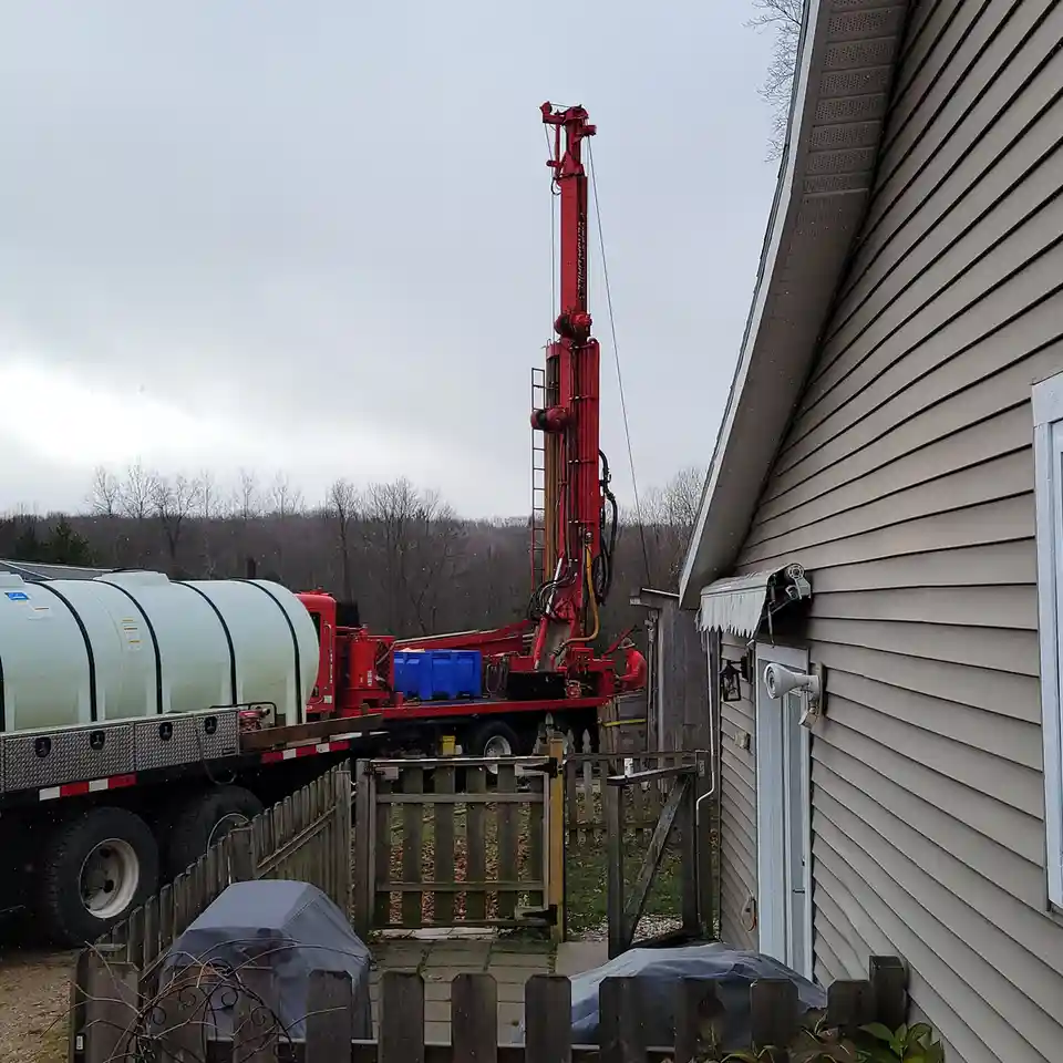 A large red water drilling rig is parked in a residential backyard next to a house, with a worker in orange gear operating…