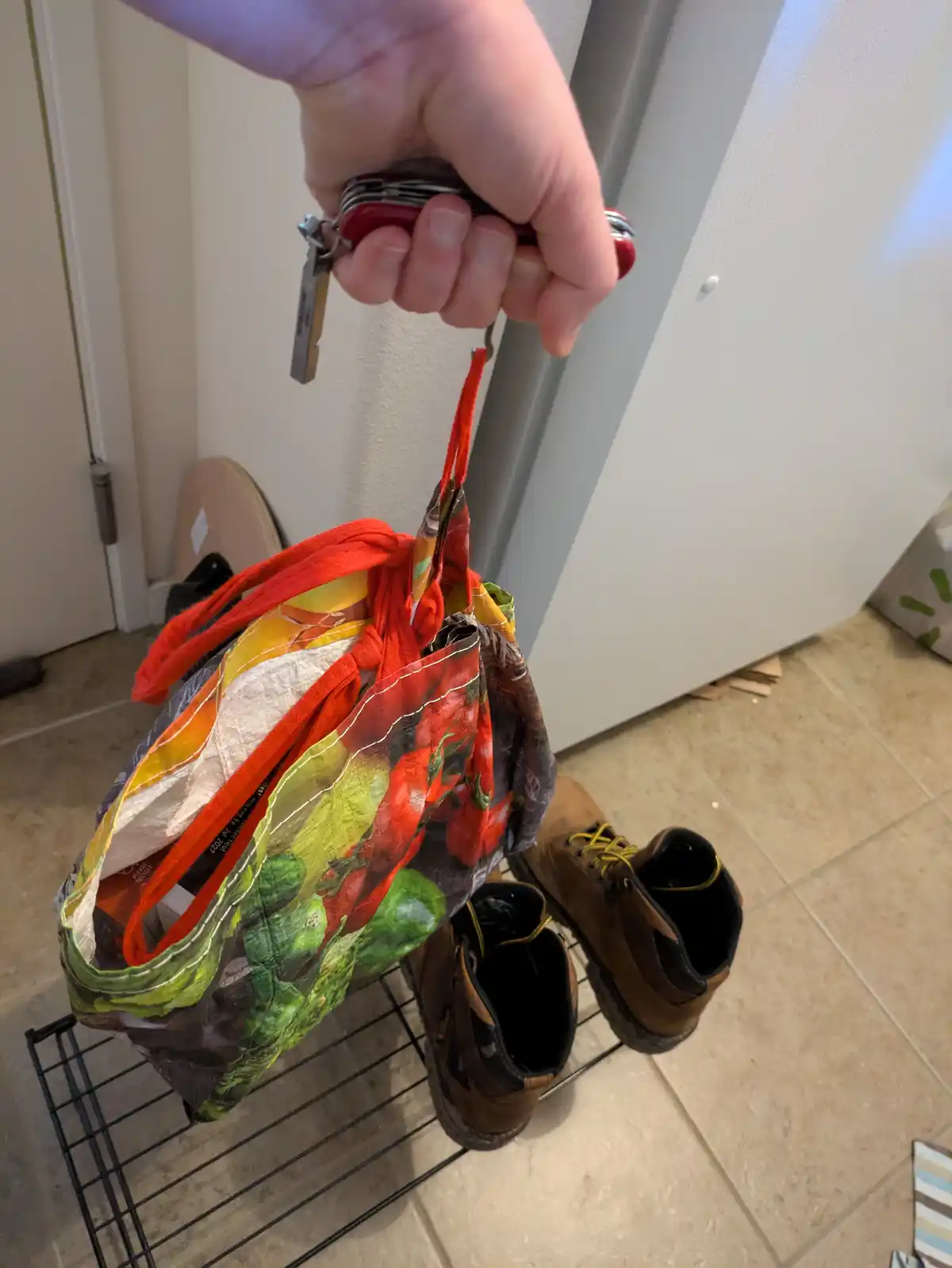 A hand grips a red Swiss Army knife attached to a keychain, holding up a colorful grocery bag printed with peppers and other…
