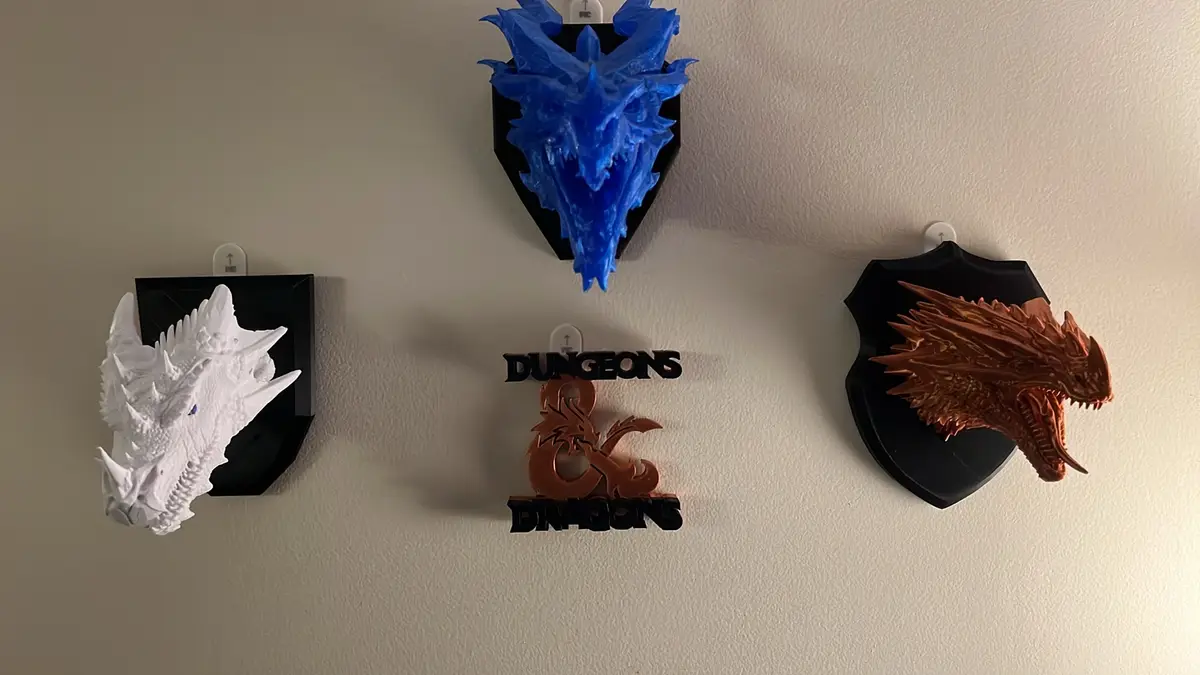 Three 3D-printed dragon head wall mounts in white, blue, and copper are displayed on a beige wall, flanking a central…