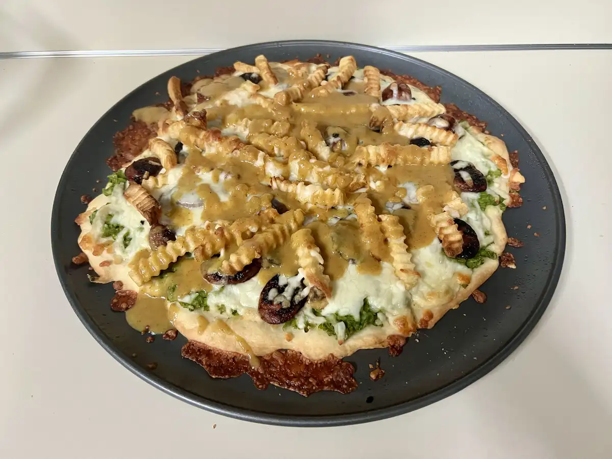 A close-up photo shows a round pizza on a dark gray plate, topped with melted white cheese, crinkle-cut french fries, a…