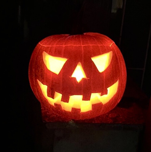 A carved pumpkin with glowing triangular eyes, a triangular nose, and a jagged toothy grin, illuminated from within against…