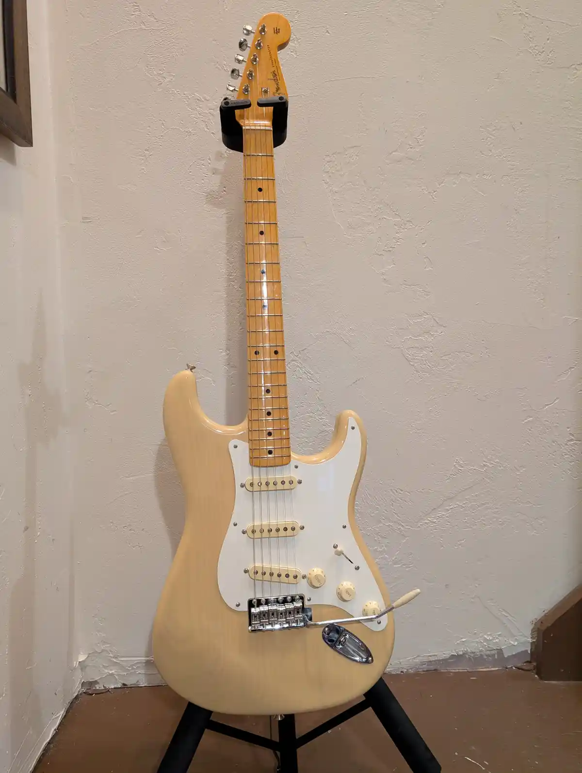 A light-wood Fender Stratocaster electric guitar stands upright on a black stand against a textured off-white wall. The…