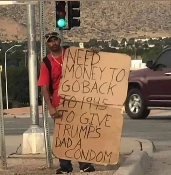 A man in a red shirt and cap stands on a street corner holding a handmade cardboard sign with handwritten text. A green…