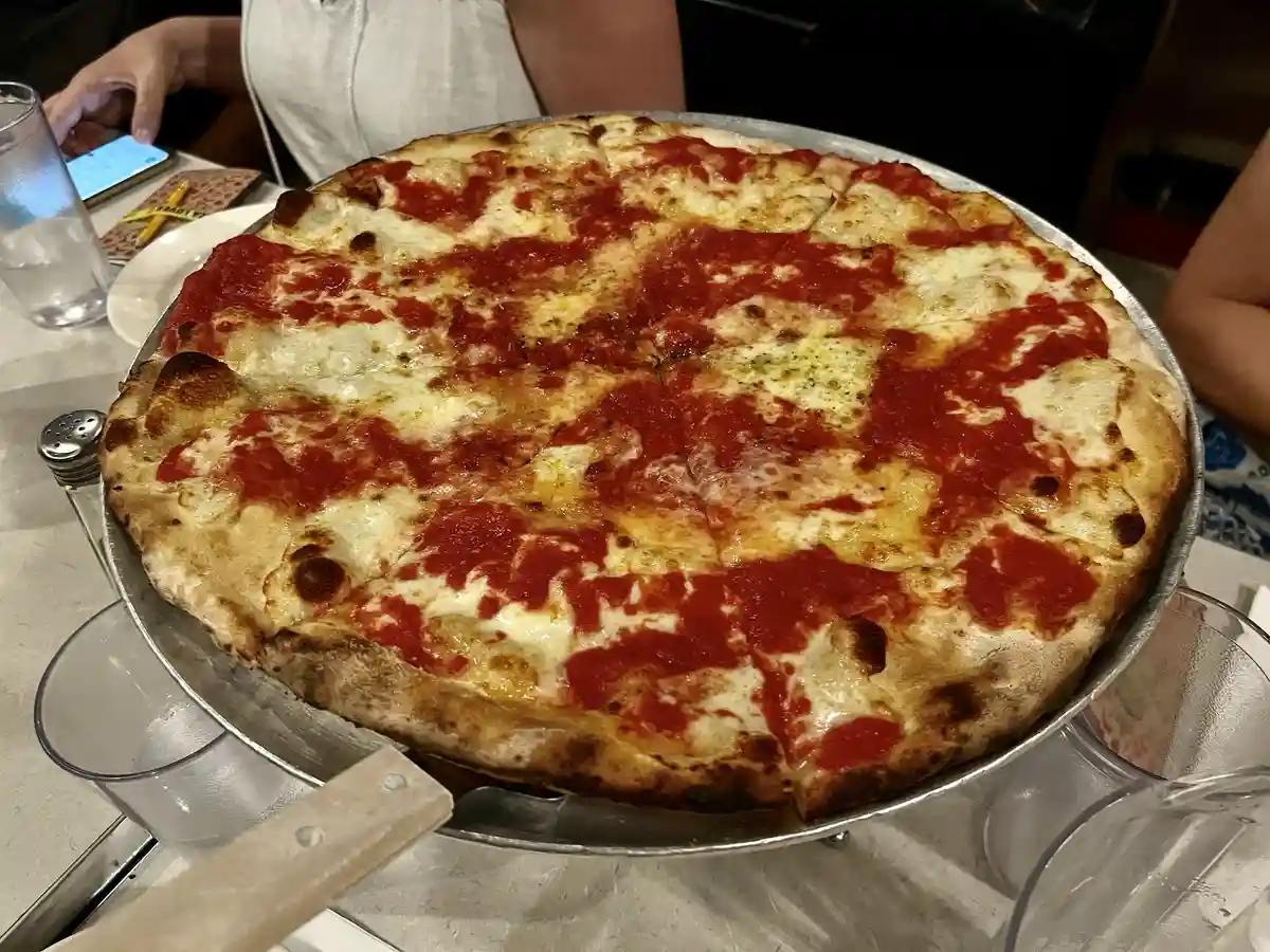 A large, freshly baked pizza with melted cheese and tomato sauce sits on a metal tray on a restaurant table, surrounded by…