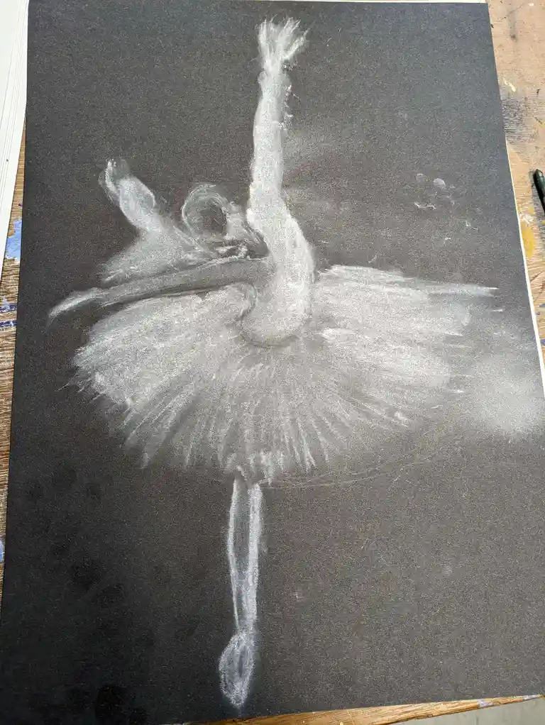 A chalk drawing on black paper depicts a ballerina in a classic pose with one leg extended and arms raised, rendered in…