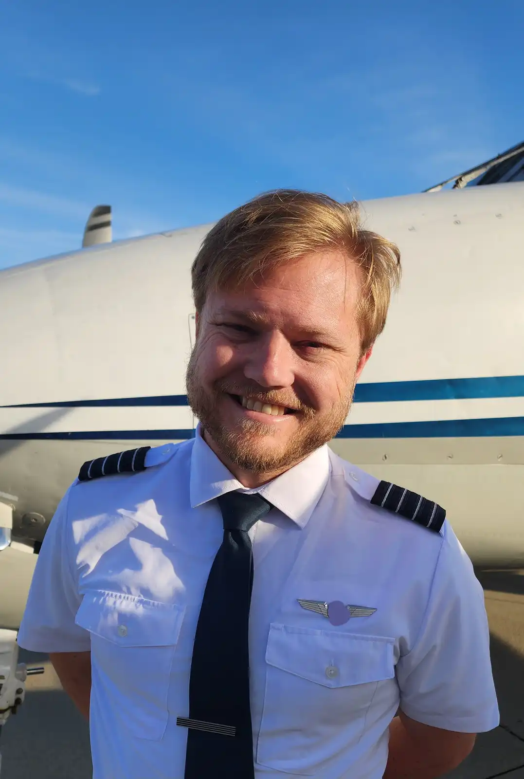 A smiling man with reddish-blond hair and a beard, wearing a white pilot uniform with epaulets and a winged pilot badge…