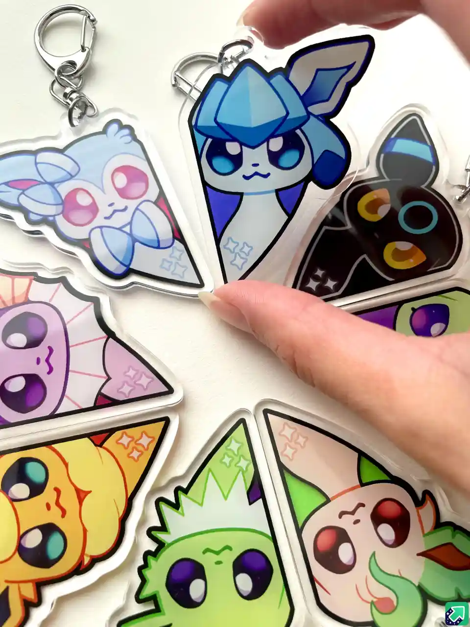 A hand holds a collection of seven colorful, chibi-style Pokémon keychains arranged in a circle on a white surface. Each…