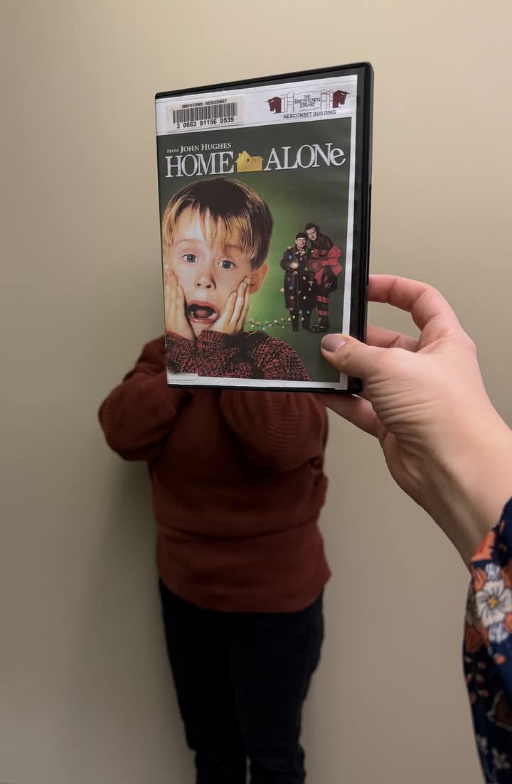 A person holds up a 'Home Alone' DVD case in front of a child, obscuring their face, so the child's shocked expression…