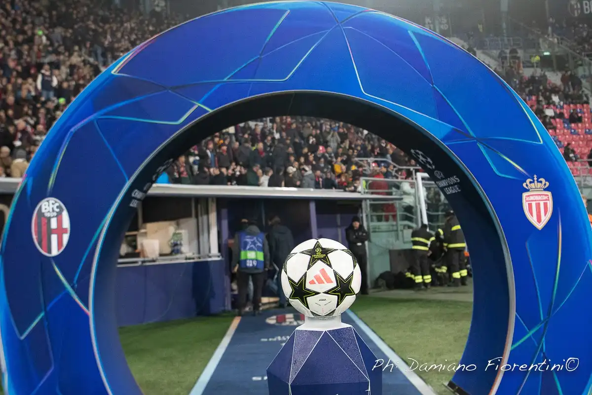 A white and black Champions League football sits atop a blue pedestal in the center of a blue tunnel archway, with the logos…
