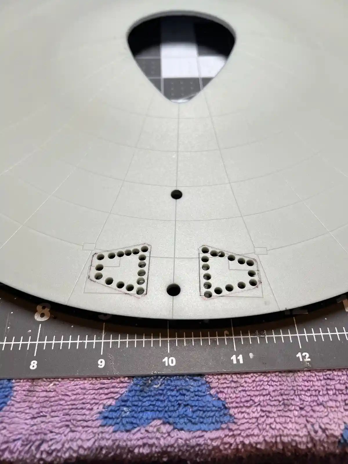A close-up photo shows two irregularly shaped, hole-punched plastic templates placed on a circular, grid-lined 3D printer…
